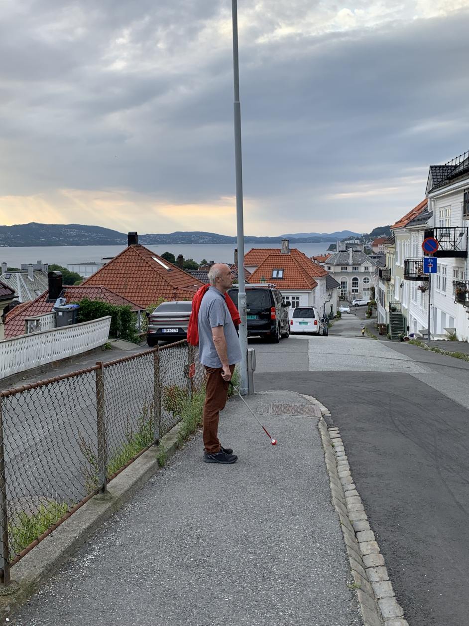 Ein Mann steht mit einem Langstock in der Hand nahe der Bordsteinkante. Er blickt auf die Straße.